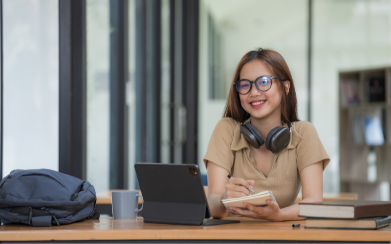 female student taking notes from a book at a library
