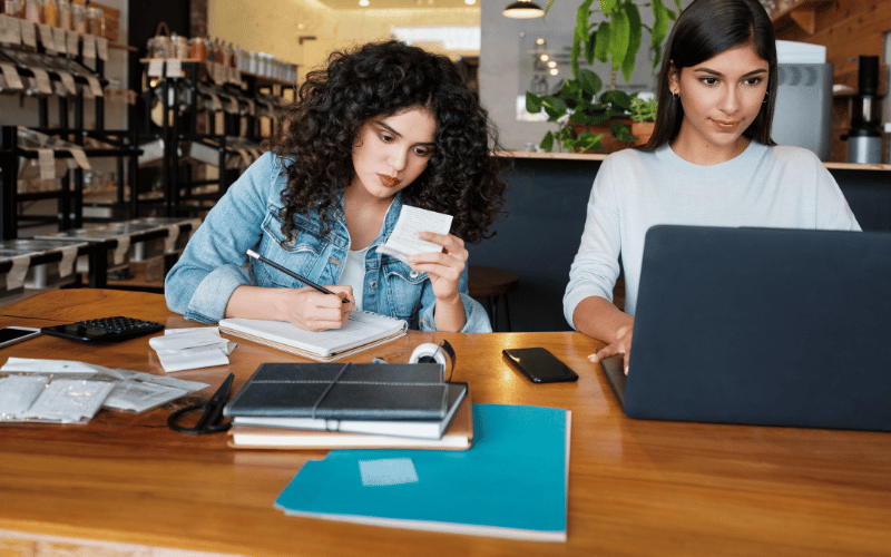 Two women working at a table, one writing and the other on a laptop