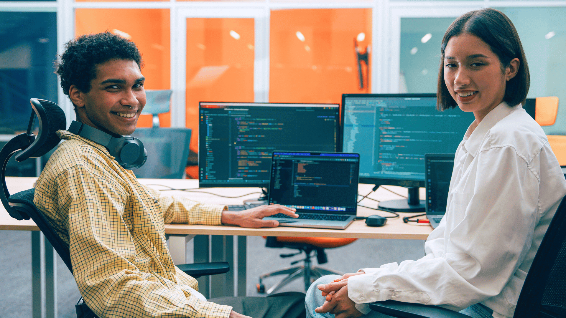 Young man and woman smile at the camera with laptops showing coding background