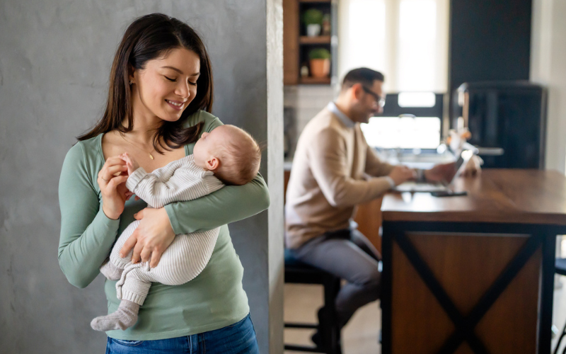 Woman cradles baby in foreground while man works in background