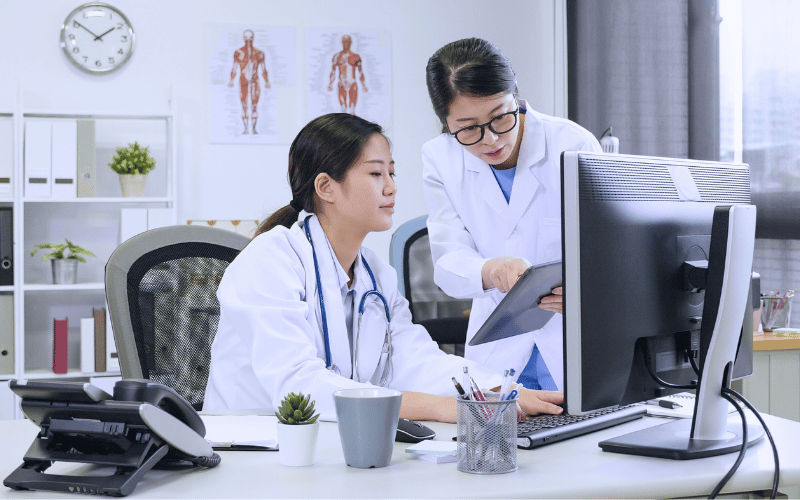 Two doctors reviewing a patient file together at a desk, working with electronic medical records on a computer.