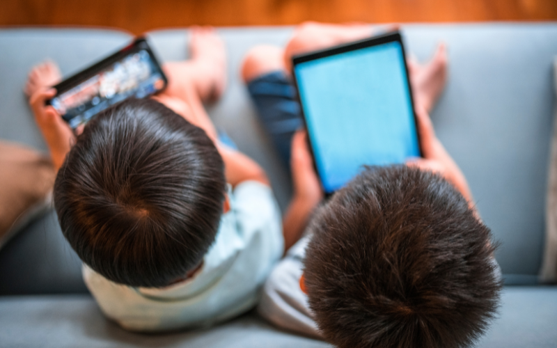 Two young children sit side by side on a couch, each absorbed in their own screen