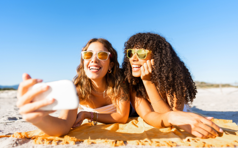 Two women wearing sunglasses lying on a beach towel, smiling, and taking a selfie