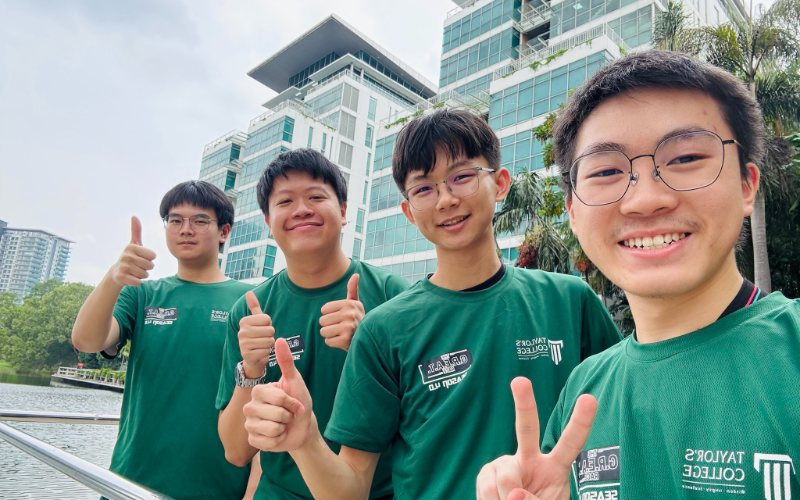 Four Taylor's College students in green Taylor's College T-shirts giving thumbs-up and peace signs outdoors on campus