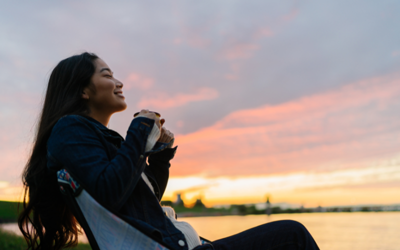 A woman sitting beside a body of water at sunset, holding a cup.