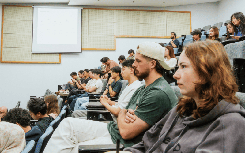 Students attending a university lecture, listening to a lecturer in a classroom.
