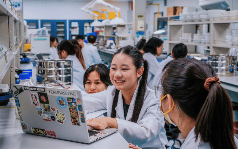 Students collaborating in a science laboratory, discussing ideas around a laptop during a practical session.