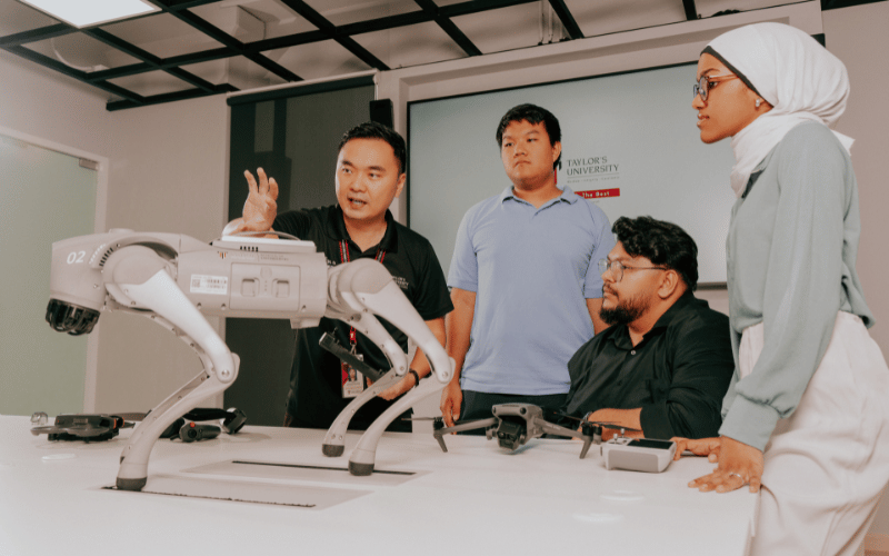 Engineering students gathered around a robotic arm and quadruped robot in a laboratory.