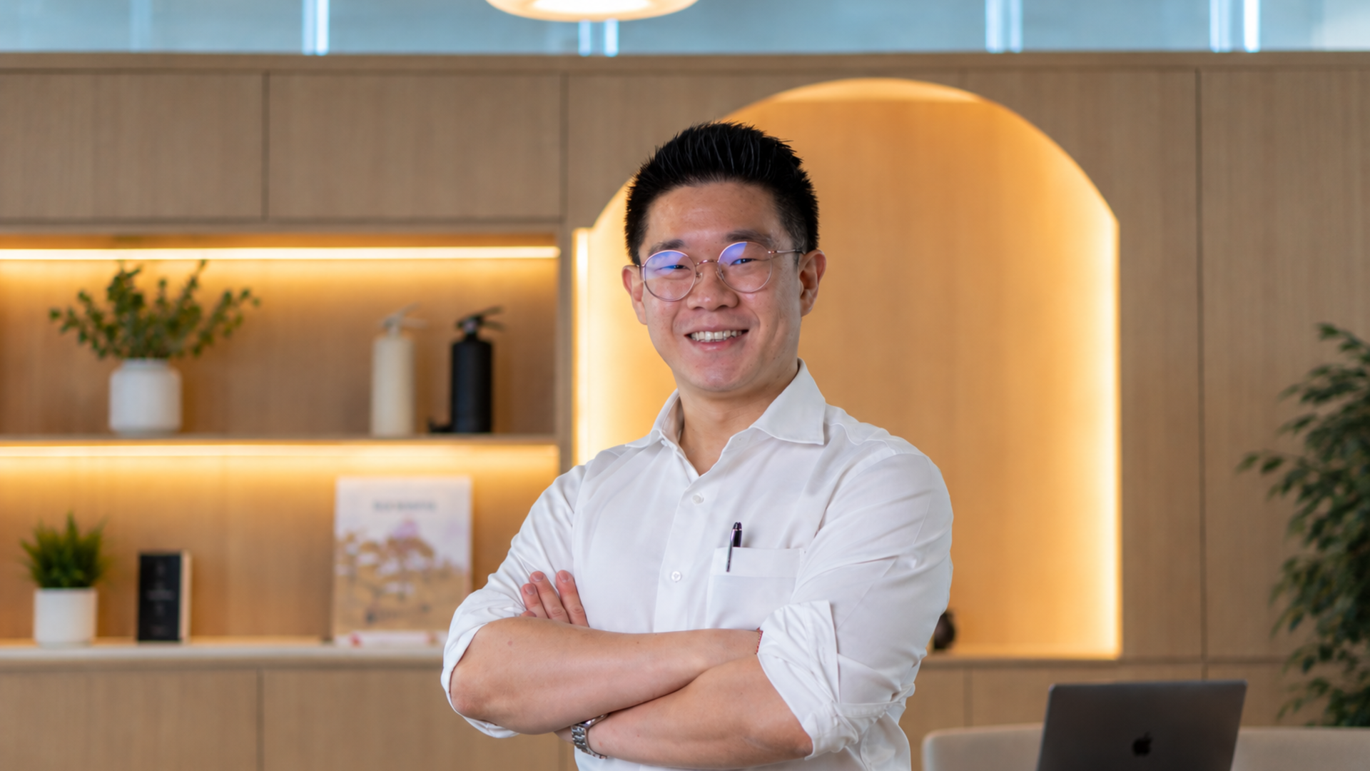Brian Soo in a white shirt, smiling with his arms crossed in front of warmly lit wooden shelving