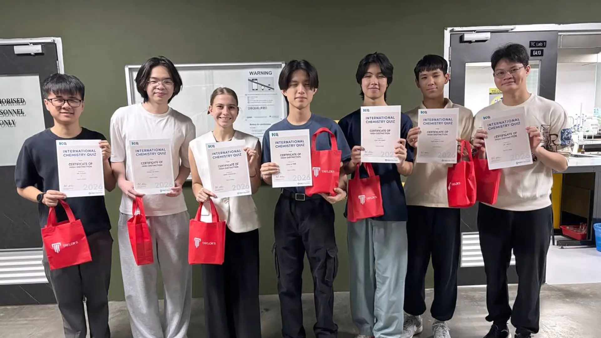 Seven people standing in a row, holding International Chemistry Quiz certificates and red Taylor's bags.