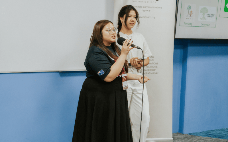 Two women giving a presentation in a classroom, with one holding a microphone while the other looks at a phone