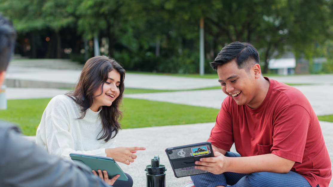 Three students sit together on the ground outdoors on campus, smiling and looking at tablets in a collaborative study session.