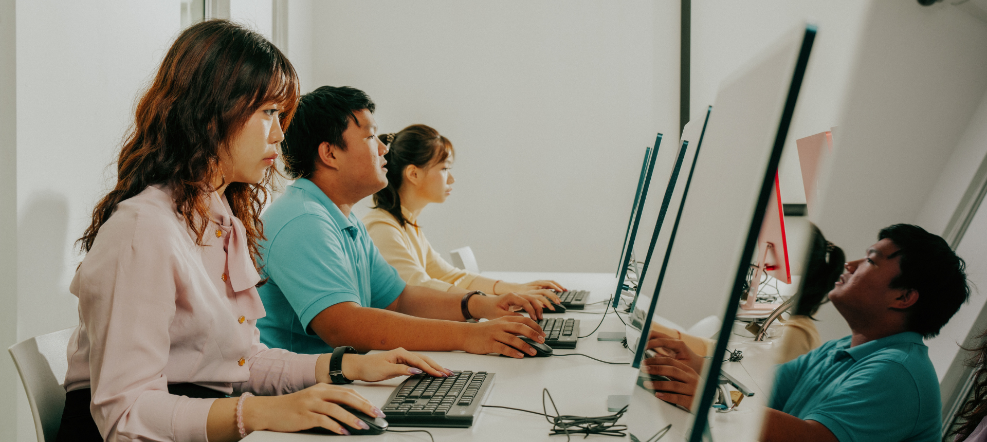 Students sit in a neat row at a long white desk, each focused on their work at a desktop computer in a brightly lit lab.