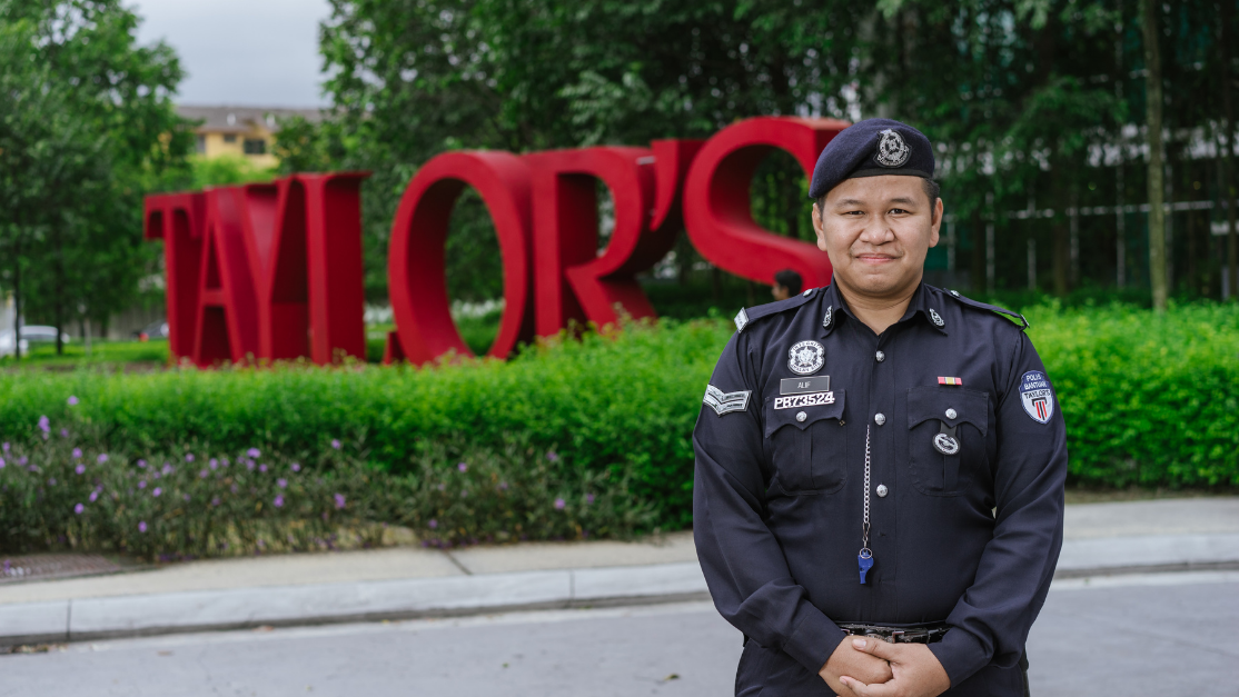 A uniformed male security guard smiles warmly at the camera while standing outdoors in front of the large, red "Taylor's" campus sign.